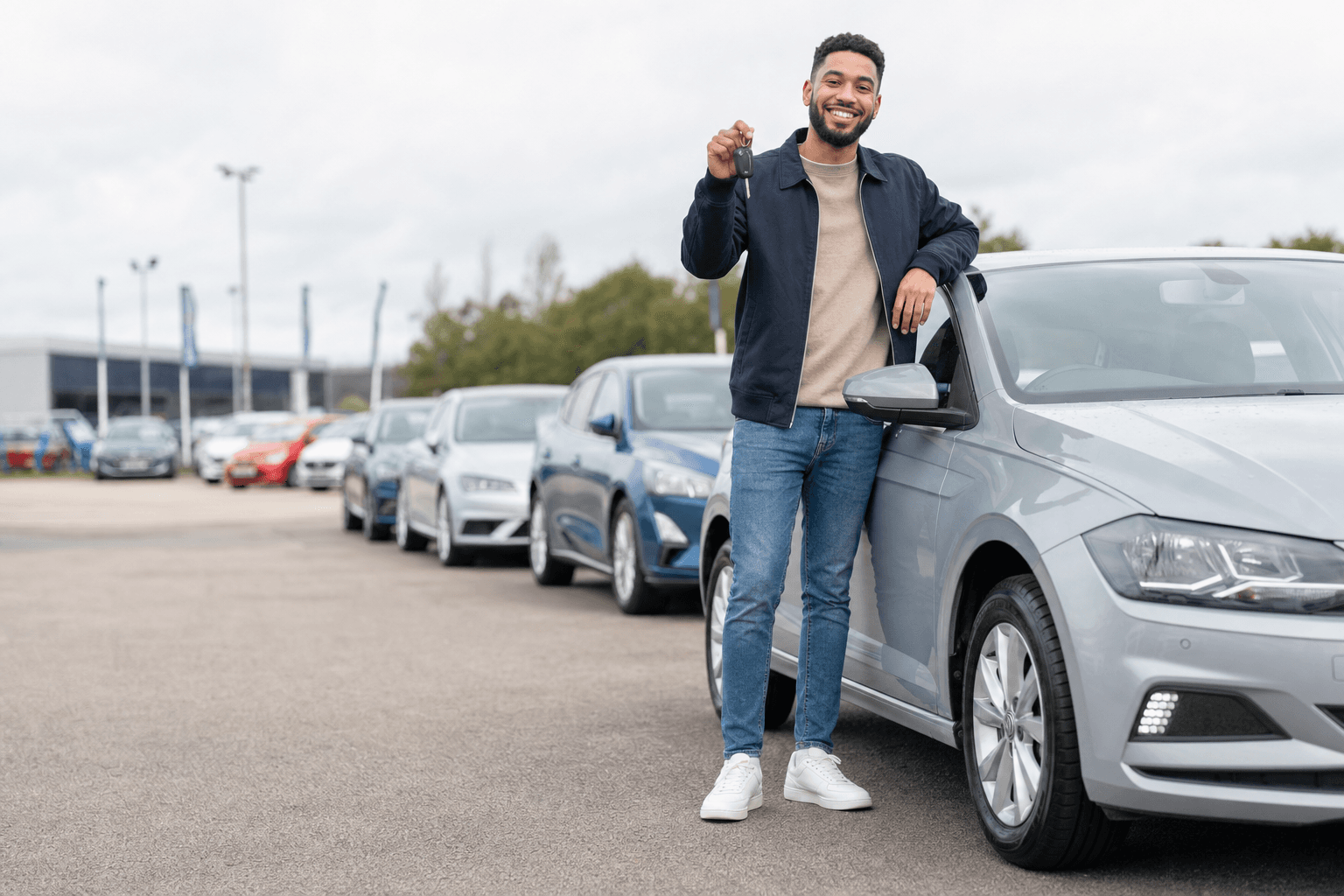 Happy customer holding car keys beside a second-hand car on a dealership forecourt